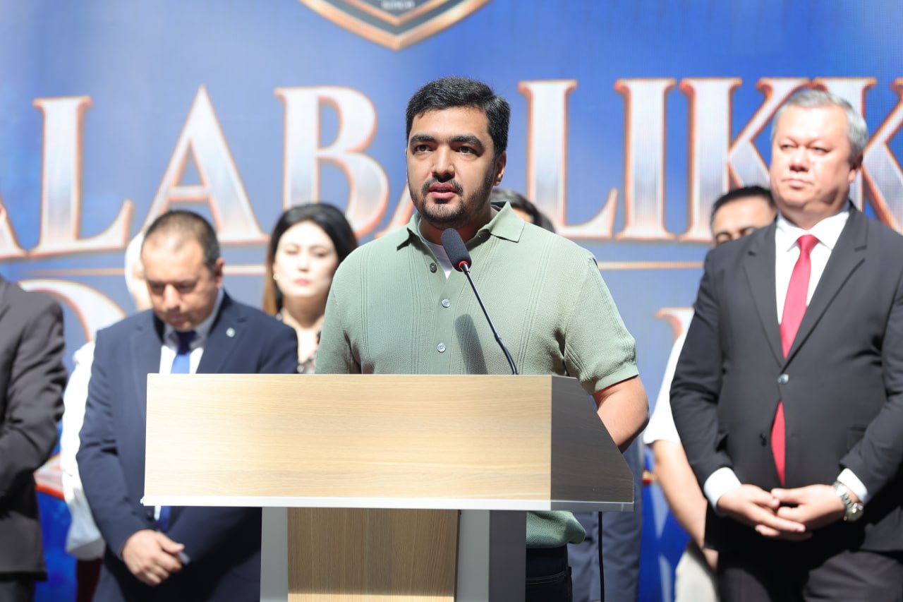 A man in a green shirt speaks at a wooden podium before a blue banner.